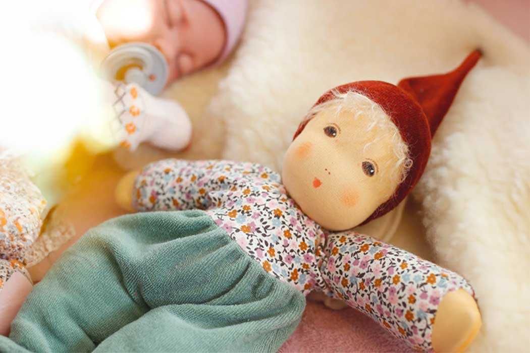 Auf dem Bild liegt ein kleines Baby auf einem weichen, hellen Untergrund. Das Baby schläft ruhig und trägt einen hellrosa Pullover. Es hat einen Schnuller im Mund und ist in einer entspannten Haltung, mit den Augen geschlossen.  Im Vordergrund liegt eine dekorative Puppe, die aussieht wie ein kleines Mädchen. Die Puppe hat ein freundliches Gesicht mit blonden, lockigen Haaren, die von einer roten, spitz zulaufenden Mütze bedeckt sind. Sie trägt ein langärmeliges Kleid mit einem bunten, floral gemusterten Stoff und grüne Stoffhosen. Das Gesicht der Puppe ist freundlich gestaltet, mit einem kleinen, zarten Lächeln und rosigen Wangen.  Das Bild vermittelt eine ruhige, liebevolle und friedliche Atmosphäre.