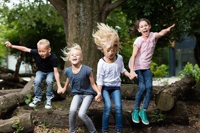 Vier Kinder springen von einem liegenden Baumstamm und halten sich dabei an den Händen. Vier Kinder springen von einem liegenden Baumstamm und halten sich dabei an den Händen.