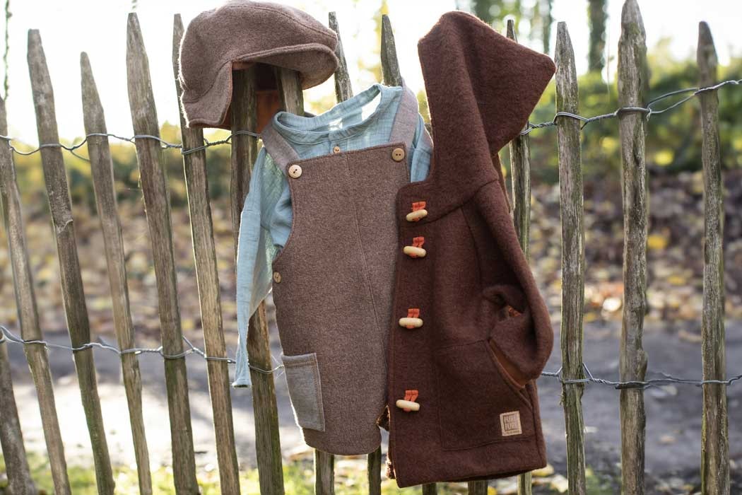 Am Zaun hängen mehrere Kleidungsstücke, die an den spitzen Holzzäunen befestigt sind. Zu sehen sind ein brauner Hut, ein hellblaues Hemd, eine braune Latzhose und eine braune Jacke mit Kapuze. Der braune Hut liegt auf einem der Zaunlatten, das Hemd hängt durch die Latzhose und die Jacke mit Kapuze ist daneben eingehängt. Die Kleidungsstücke bestehen aus warmem, weichem Stoff, wahrscheinlich Wolle oder Filz. Im Hintergrund sieht man einen natürlichen Außenbereich mit Bäumen und einem kleinen Wasserlauf. Das Licht ist hell, es wirkt wie an einem sonnigen Tag. Die Szene vermittelt den Eindruck von Herbst oder Frühling, mit einem ruhigen und ländlichen Ambiente.