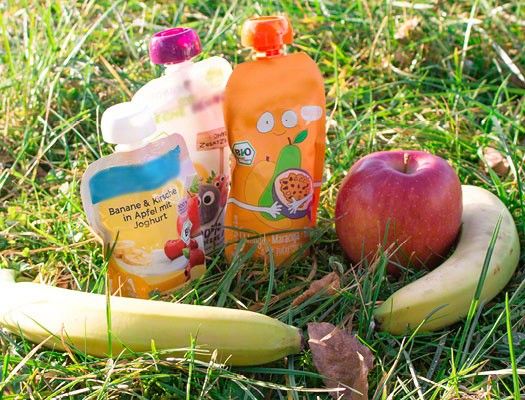 3 verschiedene Quetschies stehen neben einem Apfel und zwei Bananen auf einer Wiese