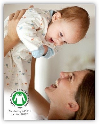 Bio Kindermode People Wear Organic Eine Frau die ein Baby hoch über Ihren Kopf hält und es anlacht. Das Baby trägt einen Schlafsack.