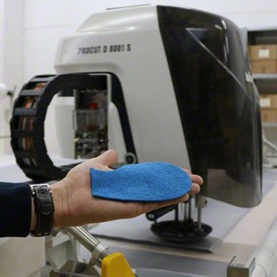 Eine moderne Schneidemaschine. Davor zeigt eine Hand ein Stück zugeschnittenes Wollwalk