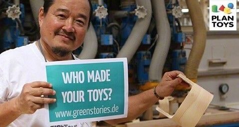 Herstellung eines Lauflernwagens bei PlanToys PlanToys-Gründer Vitool hält einen Rohling des Lauflernwagens und ein Schild mit der Aufschrift "Who made your toys?"