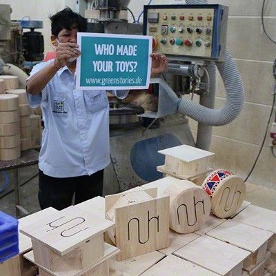 Verschiedene Stadien der Kindertrommel aus Holz bis hin zur fertigen Kindertrommel und ein Mitarbeiter der ein Schild mit der Aufschrift "Who made your toys?" hochhält