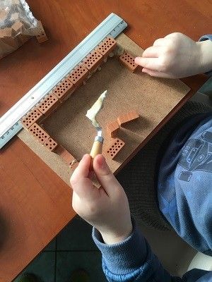 Man sieht die Hände eines Kindes von oben, wie es aus kleinen Steinen und einer kleinen Kelle voll Mörtel eine Wand baut