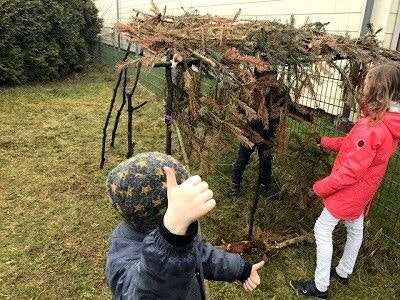 Hütten bauen mit Stick-lets Kinder mit einem fertigen "Haus" aus Ästen und Stick-lets, auf dem Dach sind Tannenzweige