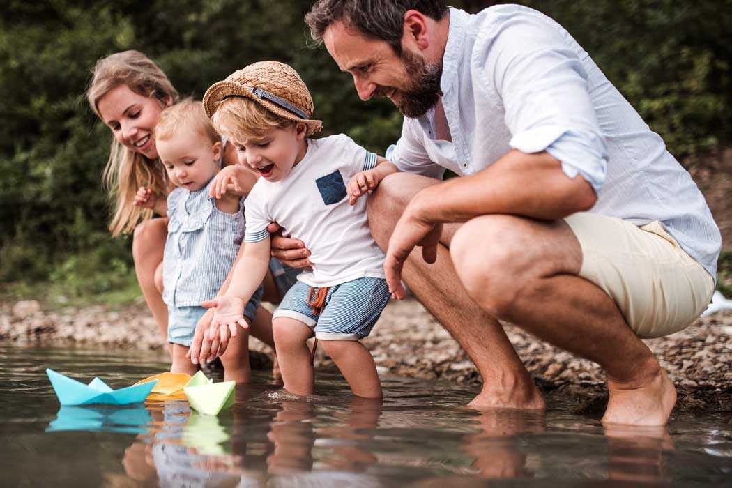 Auf dem Bild sind eine Familie mit vier Mitgliedern am Ufer eines Flusses. Sie stehen im Wasser, das bis zu ihren Knien reicht. Auf der linken Seite ist eine junge Frau mit langen, hellbraunen Haaren, die lächelnd auf die Kinder schaut. Neben ihr steht eine kleine Tochter mit blonden Haaren, die ebenfalls fröhlich aussieht. In der Mitte sind zwei kleine Jungen. Der ältere Junge trägt einen Hut und ein Kurzarmshirt mit Streifen und zeigt auf etwas im Wasser, was wie ein kleines Papierboot aussieht. Seine Mimik ist lachend und begeistert. Der jüngere Junge, der in den Armen der Erwachsenen gehalten wird, schaut neugierig auf das Wasser und die Boote darin. Auf der rechten Seite ist ein Mann mit dunklem, braunem Haar und Bart, der ebenfalls lächelnd die Kinder beobachtet und sie hält. Alle scheinen Spaß zu haben, lachen und genießen den sonnigen Tag am Wasser. Im Hintergrund sieht man Bäume und grüne Natur, die eine ruhige und friedliche Atmosphäre schaffen.