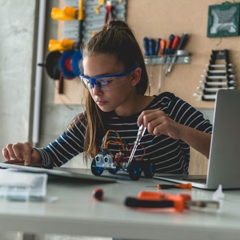 Teenagerin werkelt mit Schutzbrille auf der Nase an einem kleinen Fahrzeug und schaut sich die Anleitung dazu an.