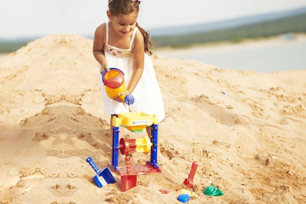 Auf dem Bild spielt ein junges Mädchen im Sand, wahrscheinlich am Strand. Sie trägt ein weißes Kleid mit dünnen Trägern und hat braunes, lockiges Haar, das zu einem Pferdeschwanz gebunden ist. Das Mädchen ist fröhlich und konzentriert sich auf ihr Spiel.  Vor ihr steht eine bunte Sandspielzeugbank mit verschiedenen Geräten und Schaufeln, die im Sand stecken oder darin liegen. Das Mädchen hält eine gelbe und rote Gießkanne in den Händen und gießt Sand in die Spielzeugbank. Im Sand um sie herum liegen weitere Spielzeuge, wie eine blaue Schaufel, eine rote Schaufel und einige kleine grüne und blaue Formen. Im Hintergrund sieht man Wasser, vermutlich das Meer, und eine weitläufige, sandige Umgebung. Die Atmosphäre wirkt sommerlich, sonnig und fröhlich.