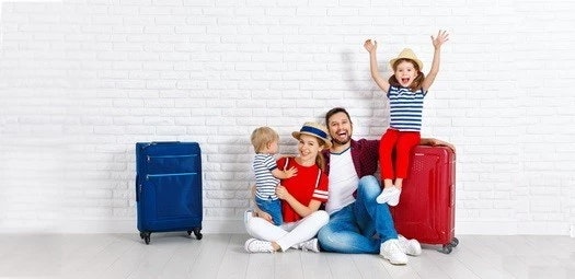 Mama und Papa sitzen auf dem Boden. Die Tochter sitzt auf einem roten Koffer und wirft die Arme in die Luft. Das kleine Geschwisterchen ist auf dem Arm der Mama. Ein blauer Koffer steht daneben.