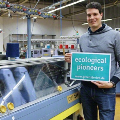 ecological pioneers disana und greenstories greenstories-Gründer Denver Mielke in den Produktionshallen von disana mit einem Schild auf dem steht "ecological pioneers".