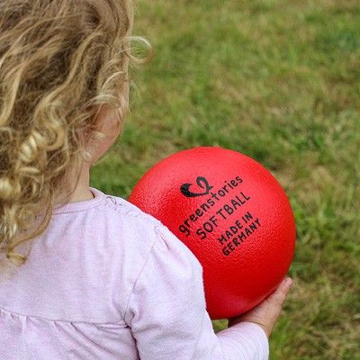 Ein Kind hält einen roten Softball in den Händen, draußen auf Rasen
