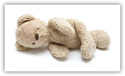 Ein liegender Teddybär vor weißem Hintergrund. Über ein Auge des Teddys sind zwei Pflaster im Kreuz geklebt.