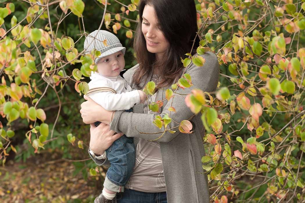 Auf dem Bild hält eine Frau ein kleines Kind in den Armen. Die Frau hat lange, dunkle Haare und trägt eine graue Jacke. Sie lächelt liebevoll und schaut auf das Kind, das sie hält. Das Kind ist ein Baby oder Kleinkind mit hellgrauem Kleidungsstück und trägt einen schicken, hellgrauen Hut mit Streifen. Es schaut neugierig und leicht ernst, während es in den Armen der Mutter sitzt. Hinter ihnen ist ein Busch mit vielen grünen und rostfarbenen Blättern, was auf eine herbstliche Stimmung hinweist. Die Szene wirkt warm, liebevoll und naturverbunden, mit den beiden im Mittelpunkt.