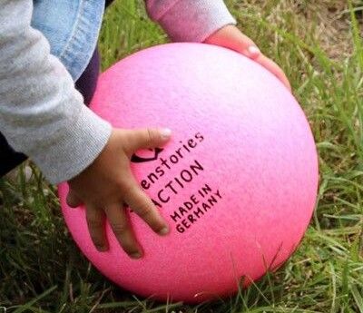 Ein pinker Ball auf Rasen, zwei Kinderhänder packen danach, das Kind ist nicht im Bild.