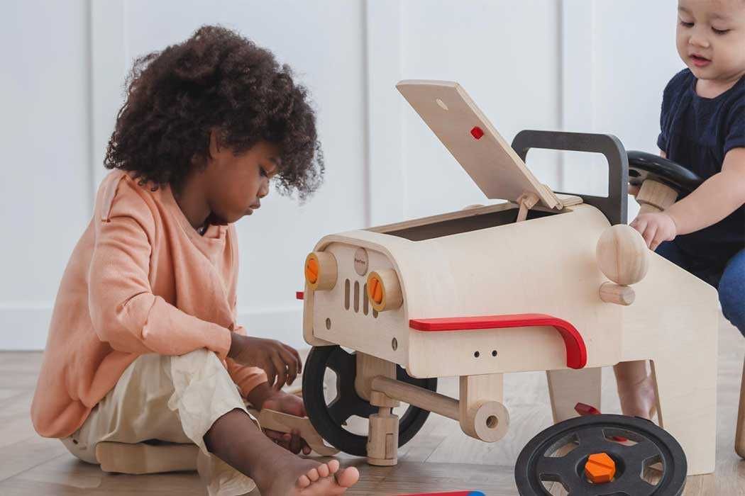 Auf dem Bild sind zwei kleine Kinder zu sehen, die mit einem Spielzeugauto spielen, das wie eine Holzpferde- oder Holzwagenform gestaltet ist. Das Spielzeugauto ist aus hellem Holz gefertigt und hat schwarze Räder mit orangefarbenen Achsen. Es hat einen kleinen roten Griff an der Seite und eine offene Klappe auf der Oberseite, die nach oben geöffnet ist.  Das Kind auf der linken Seite sitzt auf dem Boden, hat dunkle, lockige Haare und trägt einen hohen, hellen Pullover in einer hellorangefarbenen Tönung sowie helle Hosen. Es ist vertieft in eine Aktivität, vermutlich spielt es im Zusammenhang mit dem Spielzeug.  Das andere Kind sitzt auf der rechten Seite, hat kurze Haare und trägt ein dunkles Shirt. Es sitzt auf einem kleinen Sitz und hat die Hände auf das Spielzeugauto gelegt, während es zuschaut. Die Umgebung wirkt hell und modern, wahrscheinlich ein Spielzimmer oder Raum zum Spielen. Die Szene strahlt eine freundliche, ruhige Atmosphäre aus.