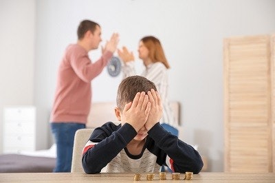Junge sitzt an einem Tisch, auf dem Geldmünzen in 5 kleinen Haufen aufgetürmt sind. Im Hintergrund streiten sich die Eltern.