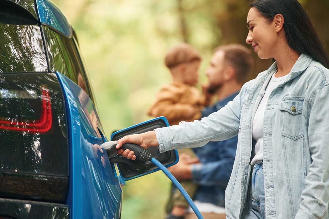 Auf dem Bild lädt eine junge Frau mit dunklen, langen Haaren ein Elektroauto auf. Sie trägt eine hellblaue Jeansjacke über einem weißen T-Shirt. Mit ihrer rechten Hand hält sie das Ladekabel, das in die Ladestation des Autos gesteckt ist. Sie steht seitlich zum Auto, schaut aufmerksam auf das Ladegerät und scheint das Laden zu überwachen.  Im Hintergrund sind zwei Männer zu sehen, die ein Kind halten. Die Männer haben kurze Haare und sind im Gespräch miteinander vertieft. Der Hintergrund zeigt eine natürliche Umgebung mit Grünflächen und Bäumen, was auf einen Park oder Wald hinweisen könnte. 