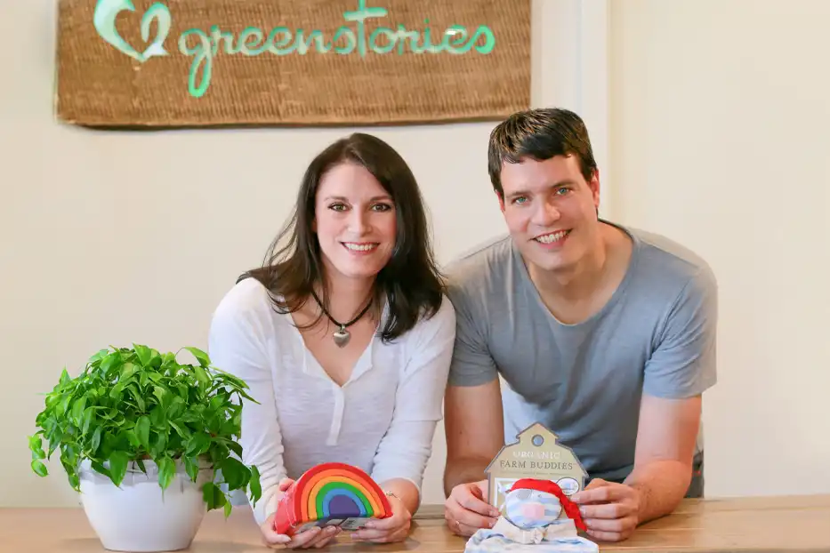 Auf dem Bild sitzen eine Frau und ein Mann an einem Tisch, die freundlich in die Kamera lächeln. Die Frau trägt ein weißes Shirt mit Knopfleiste und hat dunkle, gewellte Haare. Der Mann trägt ein graues T-Shirt und hat kurze, dunkle Haare. Zwischen ihnen liegt eine Tafel oder ein Banner im Hintergrund, auf dem in grüner Schrift „green stories“ steht; das Wort „green“ ist mit einem Herzsymbol versehen.  Auf dem Tisch vor ihnen liegt eine weiße Topfpflanze mit sattem, grünem Laub. In den Händen halten sie beide jeweils ein buntes, gewölbtes Objekt: Die Frau hält einen Regenbogen, der in schönen, kräftigen Farben gestaltet ist. Der Mann hält ein weiteres Objekt, das wie ein Miniatur-Haus mit einem Schild „Farm Buddies“ ist, darunter eine kleine, farbige Dekoration. Die Szene wirkt warm, freundlich und entspannt, mit einem Fokus auf Umwelt- oder Nachhaltigkeitsthemen.