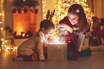 Mutter und ihre zwei Kinder sitzen auf dem Boden in einem weihnachtlich dekorierten Zimmer und schauen in eine glitzernde Box, aus der es leuchtet. Mutter und ihre zwei Kinder sitzen auf dem Boden in einem weihnachtlich dekorierten Zimmer und schauen in eine glitzernde Box, aus der es leuchtet.