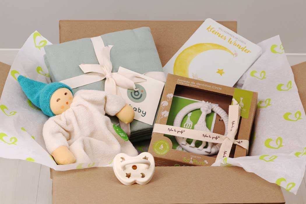 Das Bild zeigt einen offenen Karton, der mit Geschenkpapier und kleinen Geschenken für ein Baby gefüllt ist. In der Mitte liegt ein Stofftier, das ein Baby mit blonden Haaren, bla bla bla blauem Mützchen und weißem Schlafanzug darstellt. Das Stofftier hat einen freundlichen Gesichtsausdruck und liegt bequem im Karton. Neben dem Stofftier befindet sich eine hellblaue Wickeldecke mit einer weißen Schleife. Außerdem ist eine Geschenkkarte mit dem Bild eines Mondes und einem Stern, sowie der Aufschrift „kleines Wunder“ sichtbar. Es gibt auch eine offene Verpackung für eine Baby-Halskette oder -Schnullerhalter, die in einem braunen Karton mit einer transparenten Fenster ist, sowie ein weißes Lätzchen mit süßem Sternendesign. Im Vordergrund liegt auch ein kleines, weiße Schnullerkette mit einem Stern- und Mondmotiv.