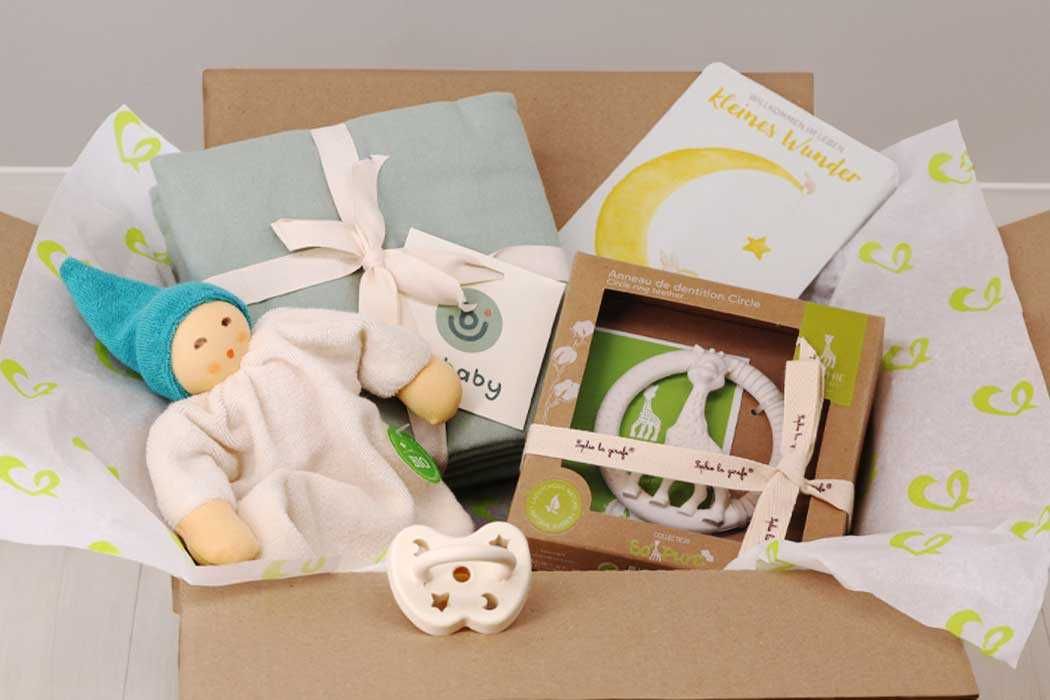 Das Bild zeigt einen offenen Karton, gefüllt mit Babygeschenken. Im Inneren liegt ein kleines, weiches Stofftier, das ein Baby mit blondem Haar, in einem weißen Schlafanzug und mit einem blauen Mützchen darstellt. Neben dem Stofftier befindet sich eine hellblaue Wickelunterlage mit weißer Schleife, sowie verschiedene Babyartikel in einer Verpackung. Darunter sind ein weiches, weißes Lätzchen mit einem Sternmuster und eine Baby-Halskette oder -Schnullerkette, die in einer braunen Verpackung mit transparentem Fenster steckt.  Der Karton ist liebevoll mit weißen Papierstreifen ausgelegt, die mit grünen Herzen bedruckt sind. 