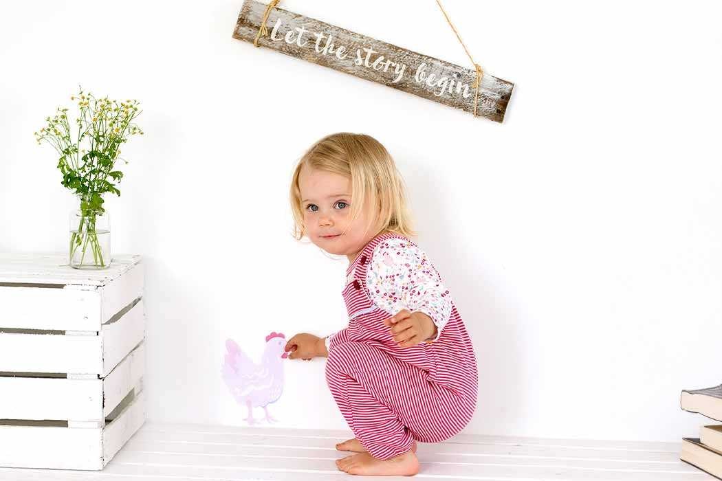 Auf dem Bild sitzt ein kleines Mädchen mit blonden Haaren auf dem Boden vor einer weißen Wand. Sie trägt einen rosa und weißen gestreiften Overall mit einem weißen Oberteil, das Muster aus kleinen roten und pinken Blumen hat. Das Mädchen schaut neugierig und leicht schüchtern, während sie mit ihrem Finger auf eine zweiteilige, rosa, schematische Zeichnung eines Huhns an der Wand zeigt. Neben ihr steht eine weiße Truhe oder Kiste, auf der ein Glas mit frischen Pflanzen oder Blumen steht. Über ihr hängt an der Wand eine Dekoration aus Holz, auf der steht „Let the story begin“ (auf Deutsch: „Lass die Geschichte beginnen“), und sie ist an einem Seil aufgehängt.