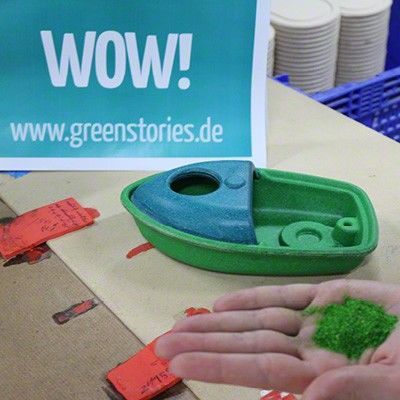 Ein Boot aus PlanWood als Badespielzeug, in grün und blau. Davor eine Hand mit grünen Farbpigmenten in der Handinnenfläche. Im Hintergrund ein Schild mit der Aufschrift "WOW!"