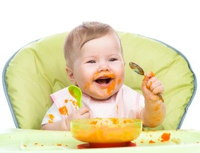 Fröhliches Baby sitz in seinem Stuhl und hat überall orangenen Babybrei