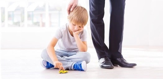 Junge sitzt gelangweilt mit einem kleinen, gelben Spielzeugauto auf dem Boden. Ein Mann in einem Anzug tätschelt ihm den Kopf.