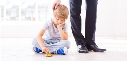 Junge sitzt gelangweilt mit einem kleinen, gelben Spielzeugauto auf dem Boden. Ein Mann in einem Anzug tätschelt ihm den Kopf.
