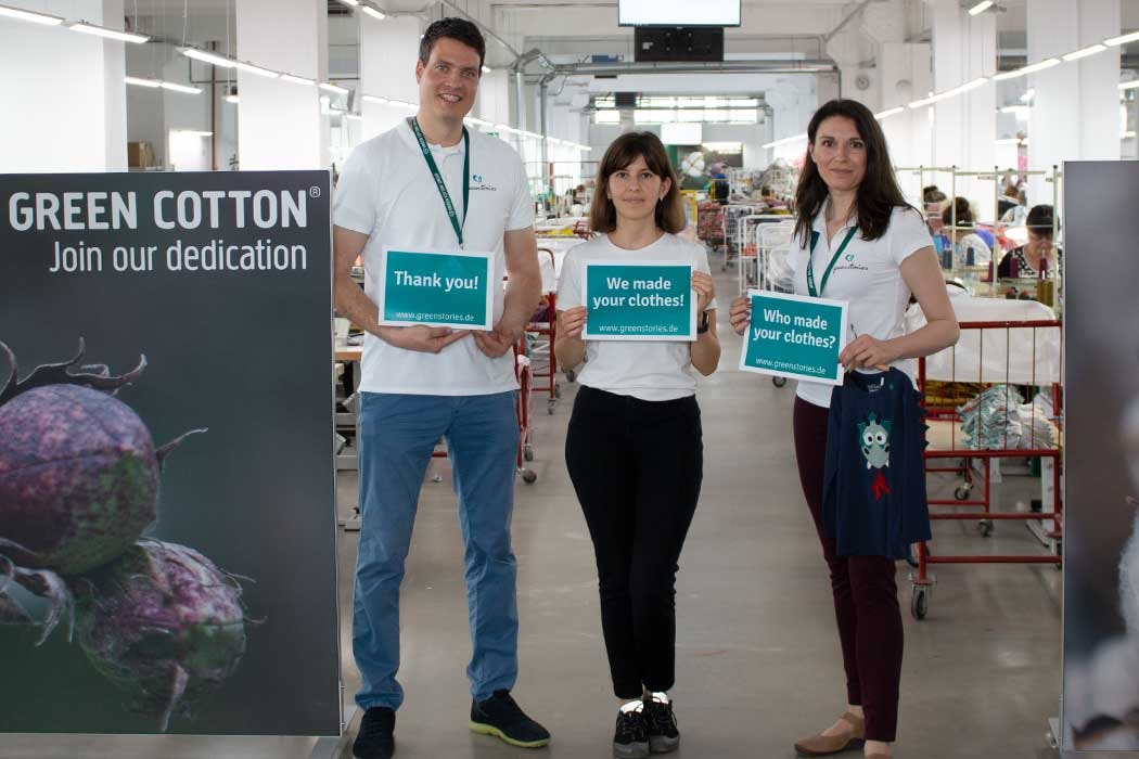 Auf dem Bild sind drei Menschen in einer Fabrikhalle oder Textilproduktion zu sehen, die vor einem großen Banner stehen. Das Banner auf der linken Seite zeigt eine große Abbildung von Baumwollfrüchten und den Text „GREEN COTTON Join our dedication“. Die drei Personen sind freundlich und lachen. Der Mann auf der linken trägt ein weißes T-Shirt mit einem Badge um den Hals und hält ein Schild, das sagt „Thank you!“ mit einer Website-Adresse. Das Mädchen in der Mitte trägt ebenfalls ein weißes T-Shirt und hält ein Schild mit der Botschaft „We made your clothes!“ und die entsprechende Website. Die Frau auf der rechten Seite trägt auch ein weißes T-Shirt, hat einen Badge um den Hals, hält ein Schild mit der Frage „Who made your clothes?“ und hält einen kleinen Kleidungsstoff mit einem lustigen, blauen Aufnäher. Hinter ihnen ist die Produktion sichtbar, mit Menschen, die an Tischen und in den Fluren der Fabrik arbeiten. Die Szene vermittelt das Thema faire Kleidung und die Herstellung von Textilien mit Fokus auf Nachhaltigkeit.