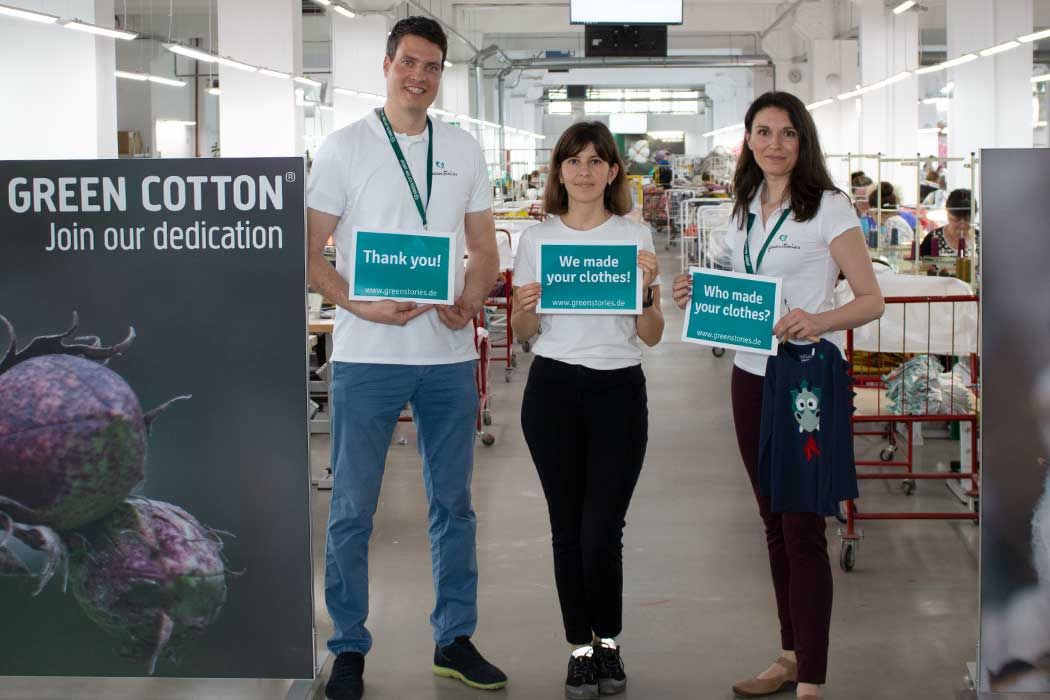 Auf dem Bild sind drei Menschen in einer Fabrikhalle oder Textilproduktion zu sehen, die vor einem großen Banner stehen. Das Banner auf der linken Seite zeigt eine große Abbildung von Baumwollfrüchten und den Text „GREEN COTTON Join our dedication“.  Die drei Personen sind freundlich und lachen. Der Mann auf der linken trägt ein weißes T-Shirt mit einem Badge um den Hals und hält ein Schild, das sagt „Thank you!“ mit einer Website-Adresse. Das Mädchen in der Mitte trägt ebenfalls ein weißes T-Shirt und hält ein Schild mit der Botschaft „We made your clothes!“ und die entsprechende Website. Die Frau auf der rechten Seite trägt auch ein weißes T-Shirt, hat einen Badge um den Hals, hält ein Schild mit der Frage „Who made your clothes?“ und hält einen kleinen Kleidungsstoff mit einem lustigen, blauen Aufnäher.  Hinter ihnen ist die Produktion sichtbar, mit Menschen, die an Tischen und in den Fluren der Fabrik arbeiten. Die Szene vermittelt das Thema faire Kleidung und die Herstellung von Textilien mit Fokus auf Nachhaltigkeit.