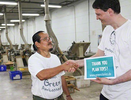 greenstories-Gründer Denver Mielke und PlanToys-CEO Vitool Viraponsavan schütteln Hände. Denver Mielke hält ein Schild in der Hand mit der Aufschrift „Thank you PlanToys!“