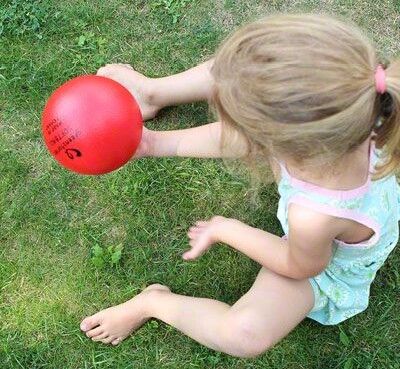 Ein Kind von oben, es sitzt auf Rasen und wirft einen kleinen roten Schaumstoffball