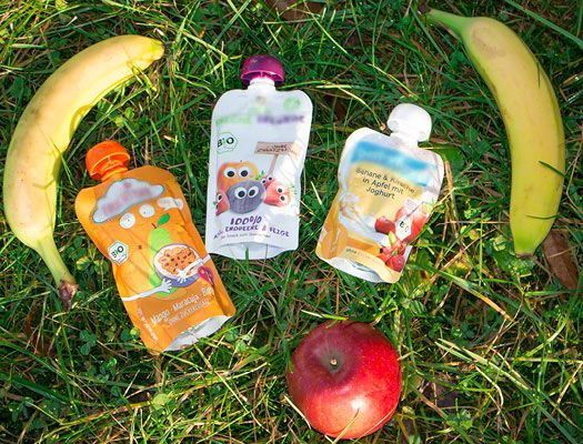 3 verschiedene Quetschies liegen neben einem Apfel und zwei Bananen auf einer Wiese
