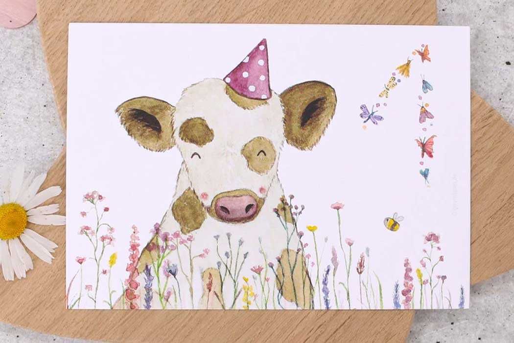 Das Bild zeigt eine fröhliche Geburtstagskarte, die auf dem Boden im Freien liegt, umgeben von grünen Pflanzen und Gras. Die Karte verfügt über eine liebevolle Illustration eines kleinen, süßen Schäfchens mit einem lachenden Gesicht und rosigen Wangen. Das Schäfchen trägt eine rosa Partyhütchen mit weißen Punkten.  In der Umgebung des Schäfchens sind kleine, zarte Pflanzen und Blumen sowie zwei summende Bienen zu erkennen. Oben rechts auf der Karte steht in einem freundlichen, gelben Schriftzug: „Alles Liebe zum Geburtstag“, wobei das Wort „Geburtstag“ hervorgehoben ist. 