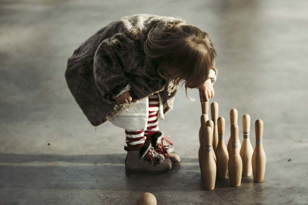 Auf dem Bild ist ein kleines Mädchen zu sehen, das gebeugt über einem Bowling-Set steht. Das Mädchen trägt einen großen, flauschigen, braunen Fake-Fur-Mantel und hat lange, braune Haare, die nach vorne hängen. Sie trägt eine weiße Hose mit roten und weißen gestreiften Stiefeln und roten Schnürsenkelschuhen. Im Moment, wo das Bild aufgenommen wurde, scheint sie gerade einen der Bowling-Kegel umzustoßen. Vor ihr stehen mehrere Bowling-Kegel, die in einer Reihe aufgestellt sind, sowie ein Bowlingball, der am Boden liegt. Das Licht ist gedämpft, und der Hintergrund ist neutral, wodurch der Fokus auf das Mädchen und die Bowling-Elemente gerichtet ist. Die Szene wirkt ruhig und spielerisch.