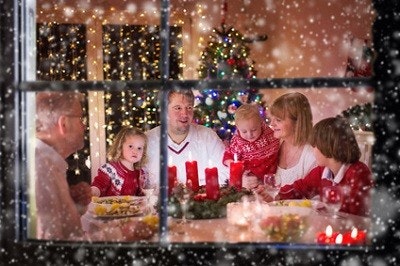 Zu Weihnachten kommt die Familie zusammen Durchs Fenster sieht man eine am Tisch zusammensitzende Familie. Ein Adventskranz steht vor ihnen, im Hintergrund ein geschmückter Weihnachtsbaum.