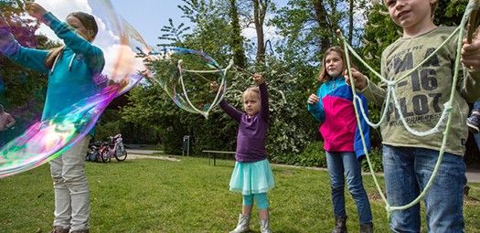 Vier Kinder stehen im Park und machen Riesenseifenblasen mithilfe von an Stäben befestigten Seilen.