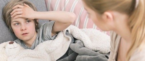 Junge liegt mit einem Teddybär auf der Couch und hält sich den Kopf, während er seine Mutter anschaut.