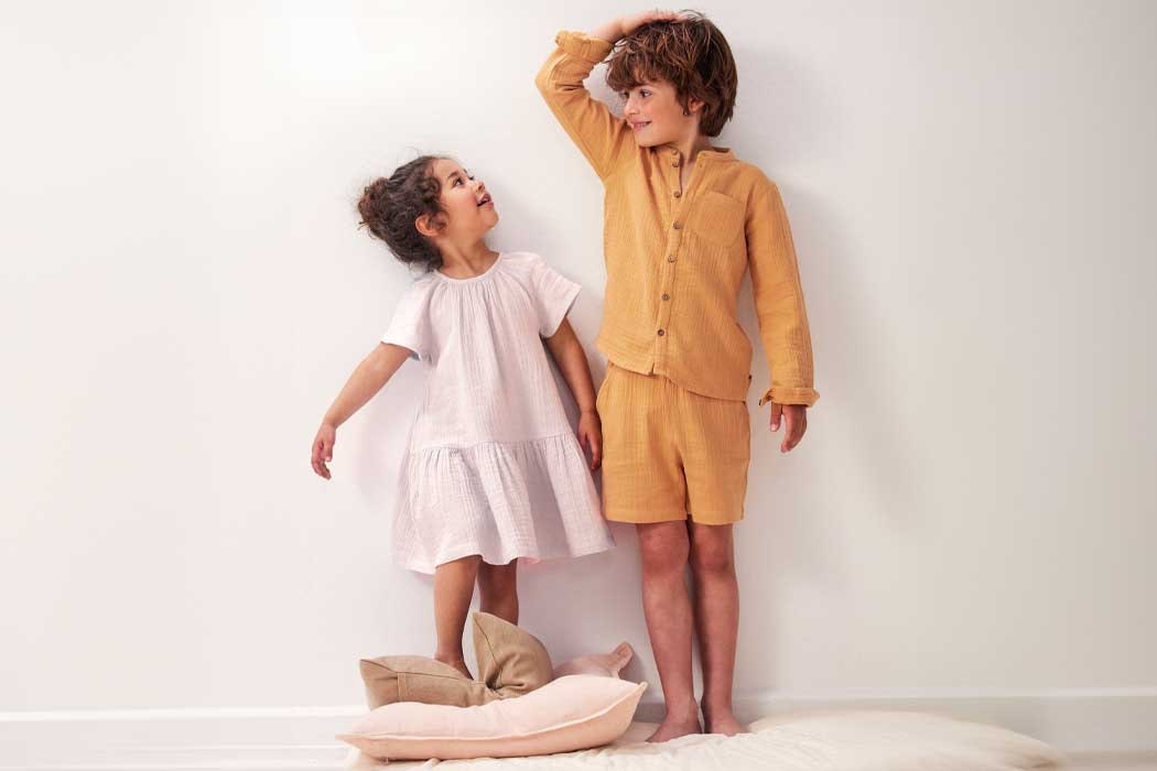 Auf dem Bild stehen zwei Kinder vor einer weißen Wand. Der Junge, der größer ist, trägt einen karamellfarbenen, lockeren Pyjama mit langer Hosen und langem Oberteil. Er hat braune, lockige Haare und ist lachend, während er mit einer Hand gegen den Kopf streicht. Das Mädchen, das kleiner ist, trägt ein helles, weißen Kleid mit kurzen Ärmeln und einer weiten, gerüschten Taille. Sie hat dunkle, lockige Haare, die zu einem Zopf zusammengebunden sind, und schaut den Jungen an mit einem fröhlichen Ausdruck. Sie haben eine innige, fröhliche Stimmung, und es sieht so aus, als würden sie miteinander lachen und Spaß haben. Das Mädchen steht auf einem Kissen, und beide Kinder stehen barfuß auf dem Boden. Die Szene wirkt warm, fröhlich und herzlich, voller kindlicher Freude